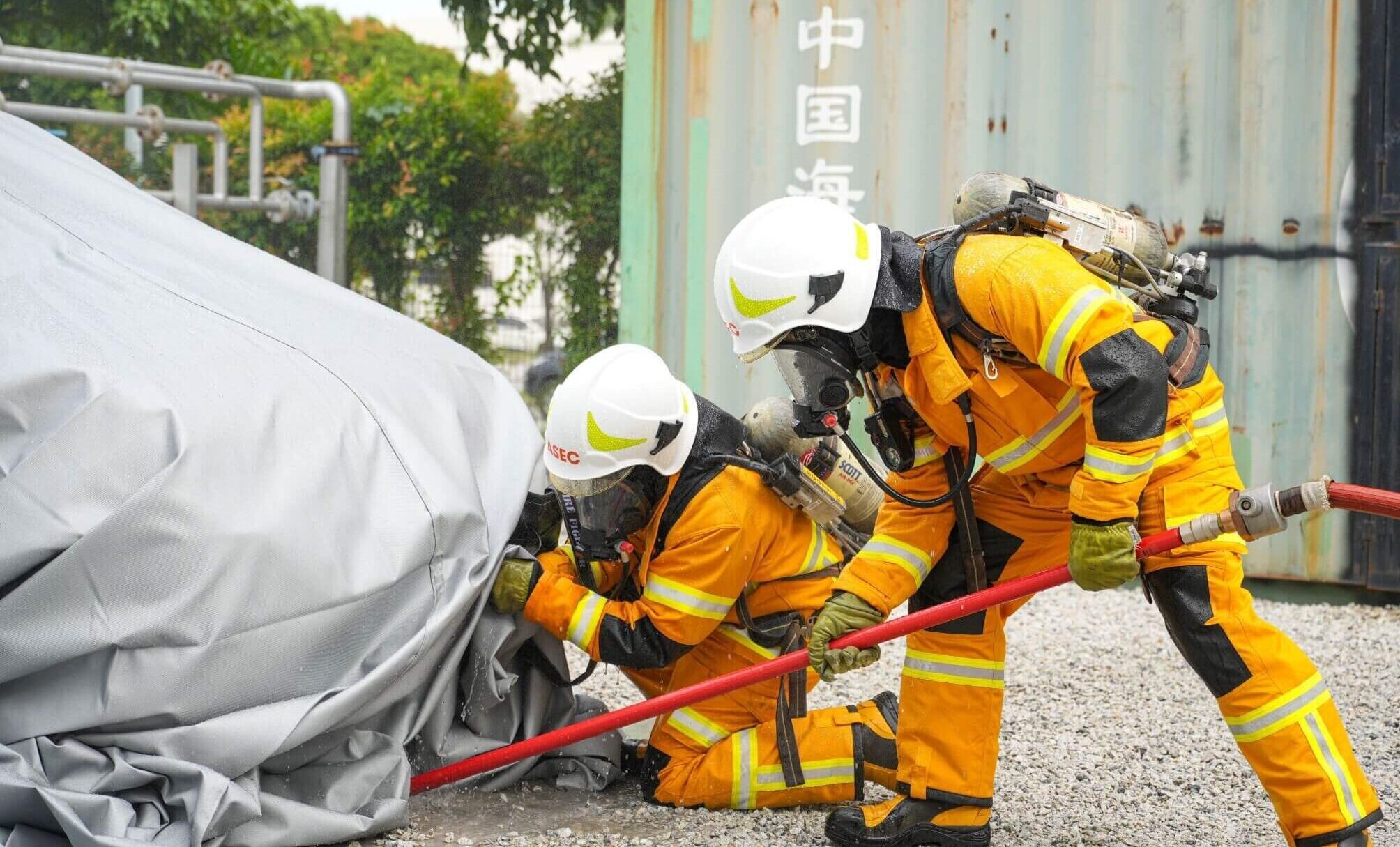 Firefighters in full protective suits performing an industrial firefighting drill with fire hoses.
