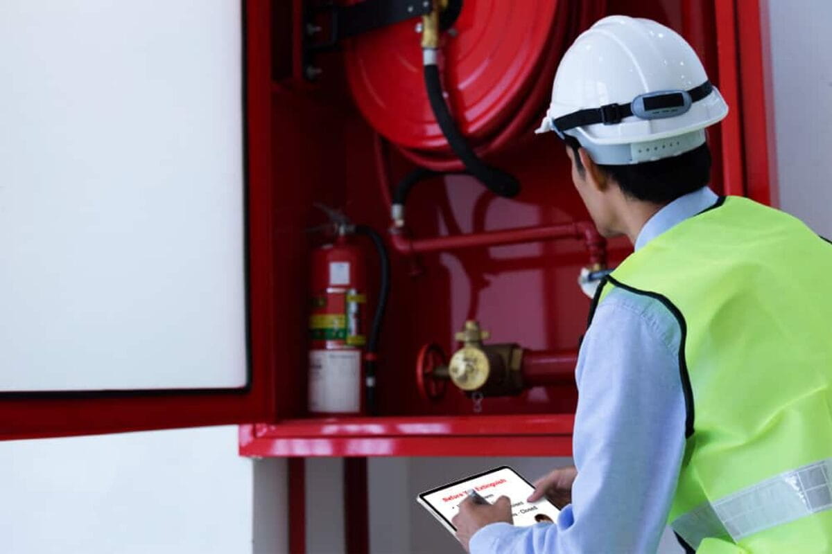 A safety inspector or technician in a yellow high-visibility vest and white hard hat examining a fire safety equipment cabinet, which contains a fire hose reel and fire extinguisher, while holding a clipboard or tablet.
